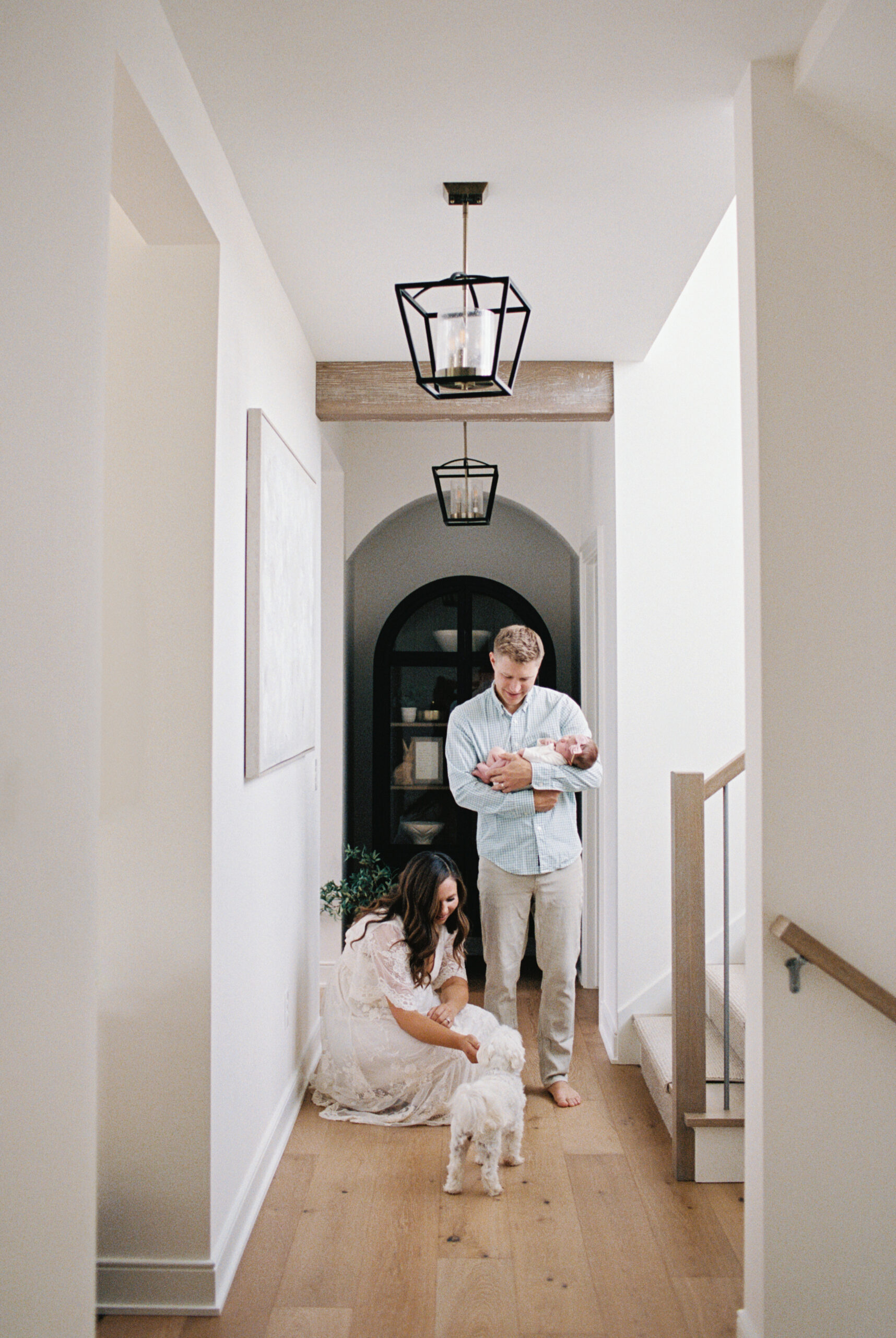 Family in long hallway, with dad in blue shirt holding onto newborn baby girl and mom in white dress kneeling down to pet small white dog