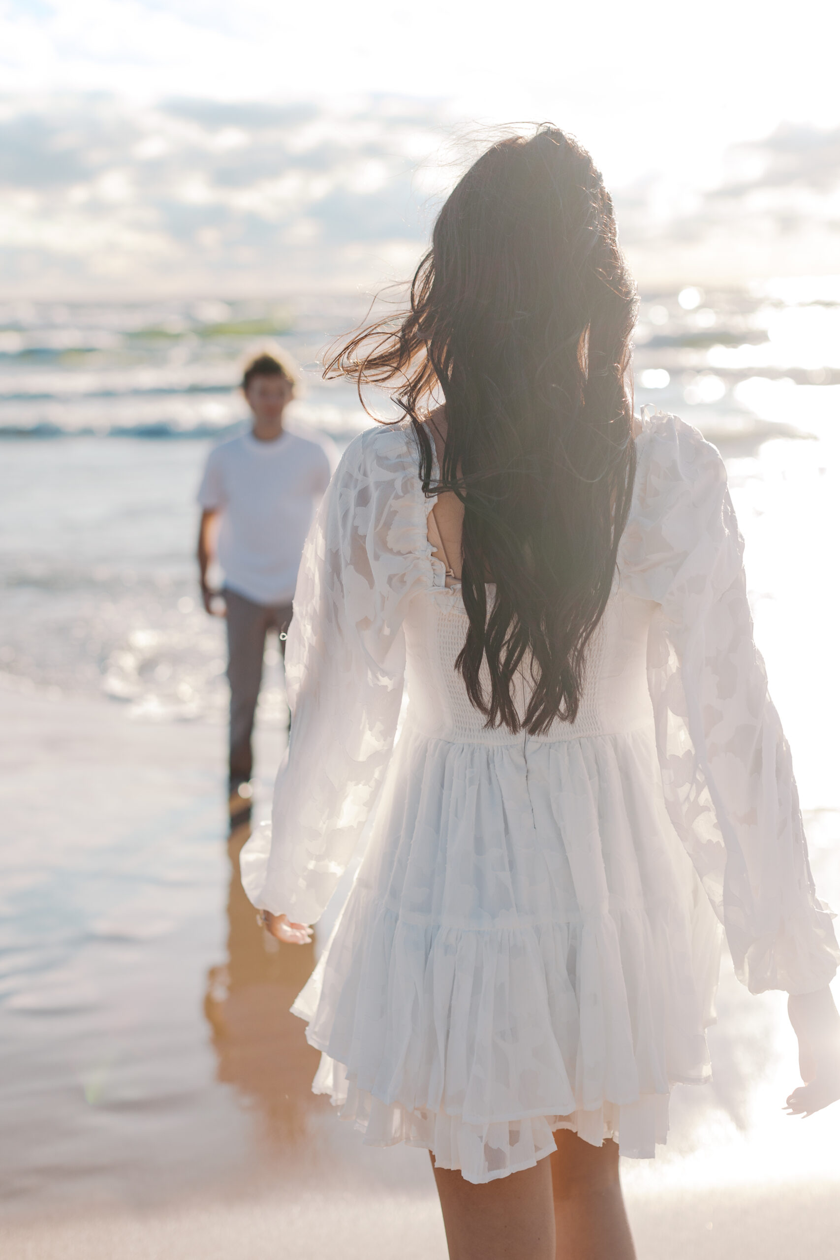 Female in white dress walking towards male in white shirt and khaki pants on a beach in Michigan with lots of sun on a very windy day with lots of waves