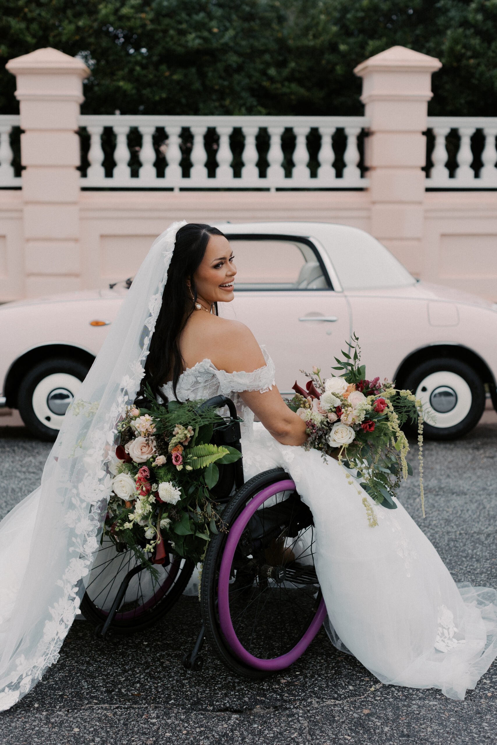 A bride in a a wheelchair with floral attached to back and bouquet on her lap in front of pink vintage car and pink and white fence in Charleston