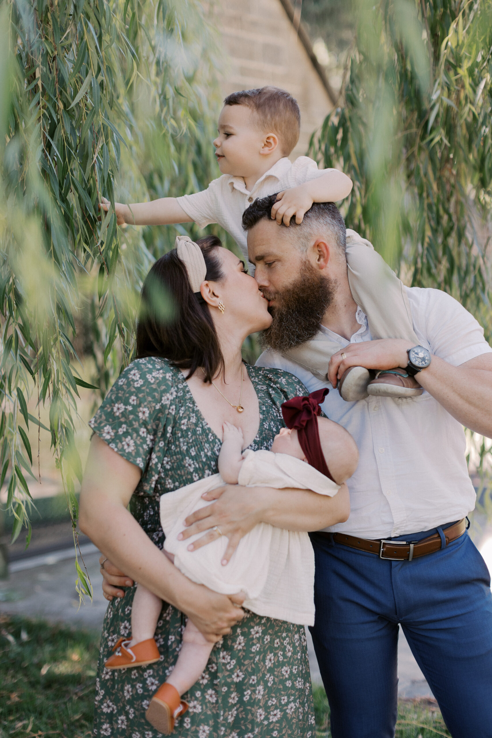 a caucasian family of four nestled inside a willow tree with mom in a green dress holding a baby girl in a white dress while dad in a white shirt has young boy on his shoulders and mom and dad lean in to share a kiss