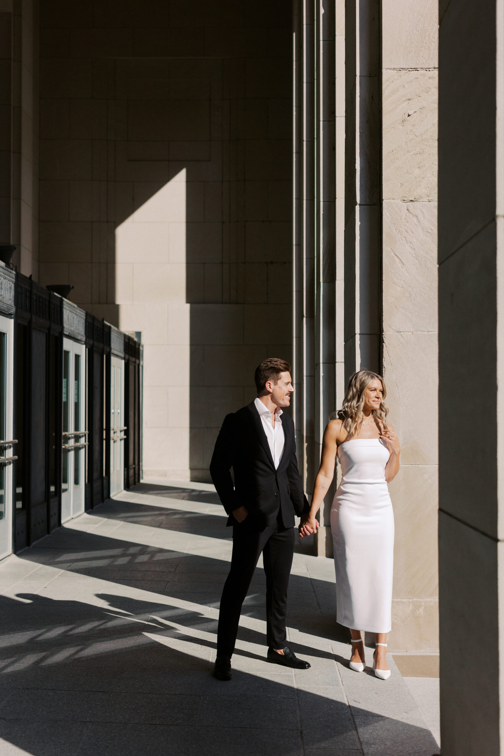 a man in a black suit and woman in a tight and short white dress holding hands with harsh shadows smiling at one another in a building with tall columns