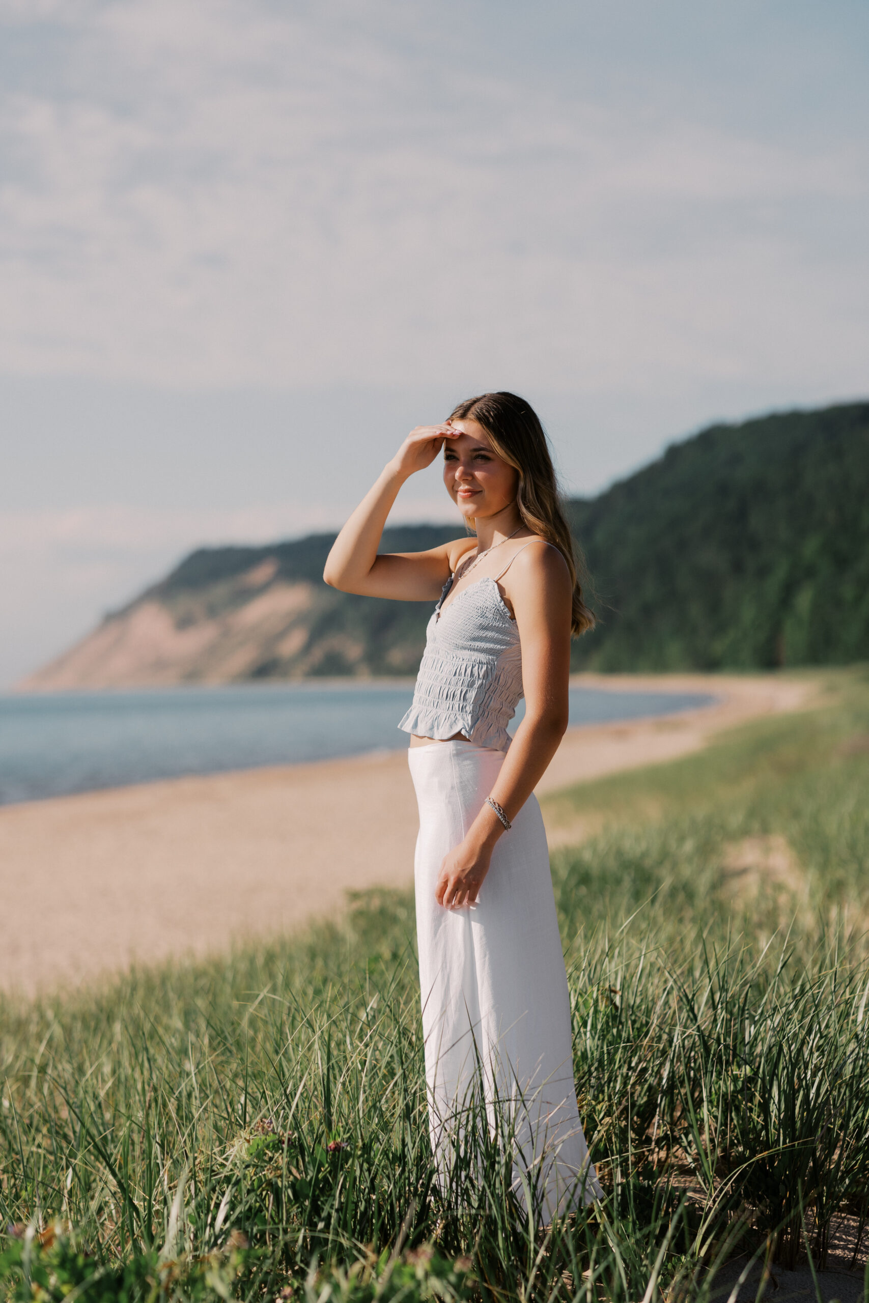 A teenage girl standing in beach grass next to a large body of water with large hills in the background wearing a white shirt and light blue tank top