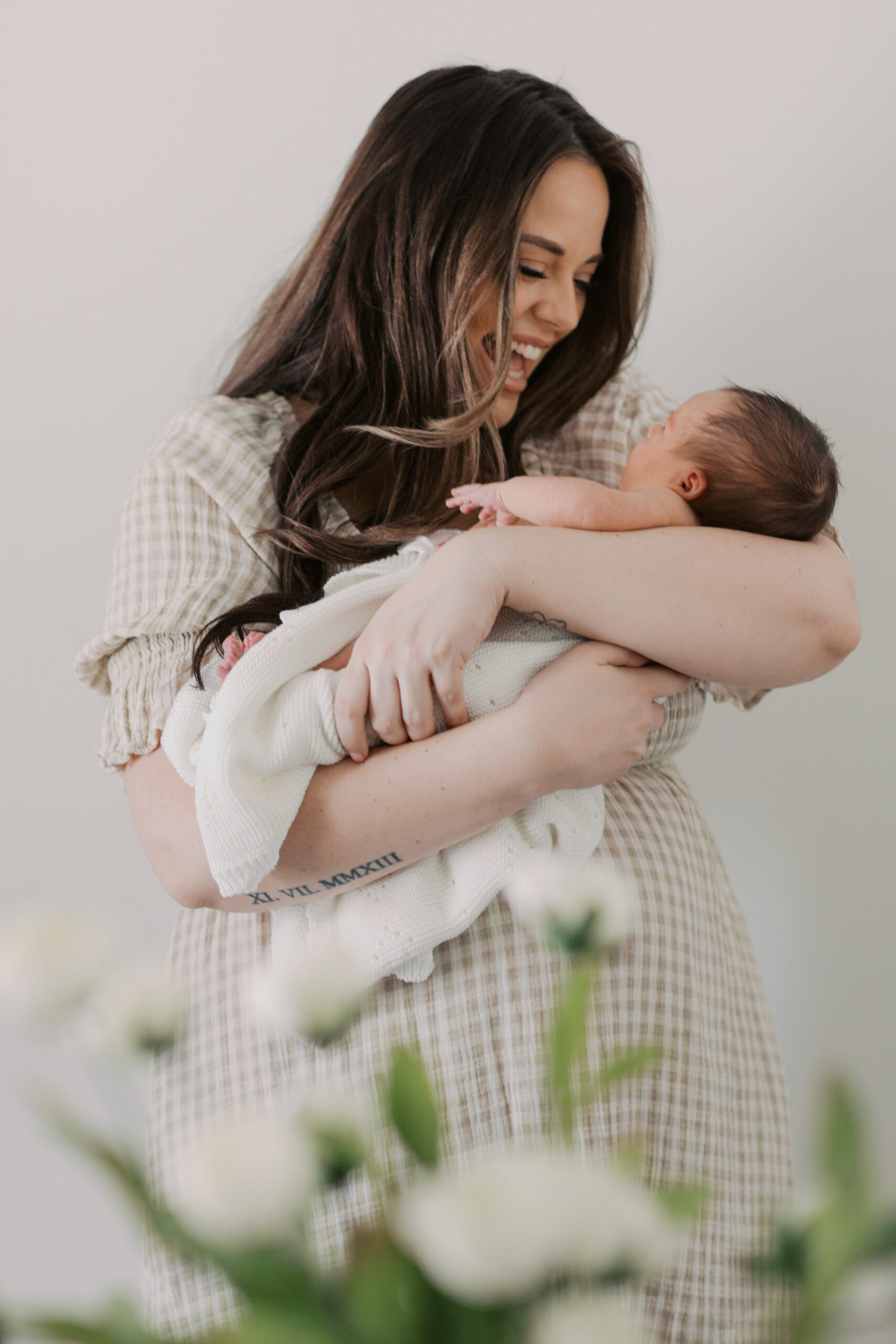 A mother with a huge smile on her face holding her newborn baby girl in her arms with flowers out of focus in the foreground