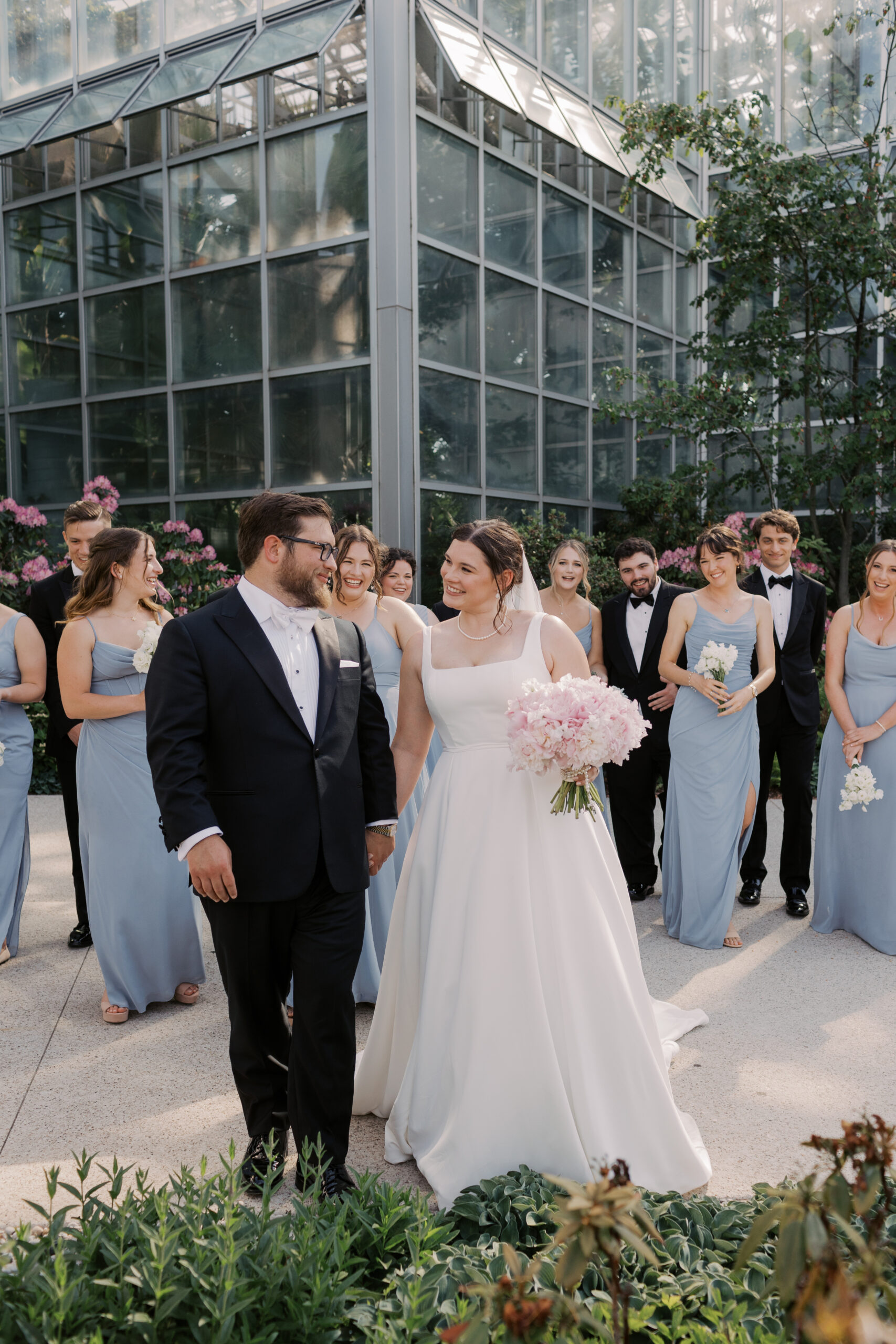 A bride with a white gown with thick straps and long cathedral veil holding the grooms hand and a bouquet of pink flowers walking toward camera in front of bridal party with happy expression on face